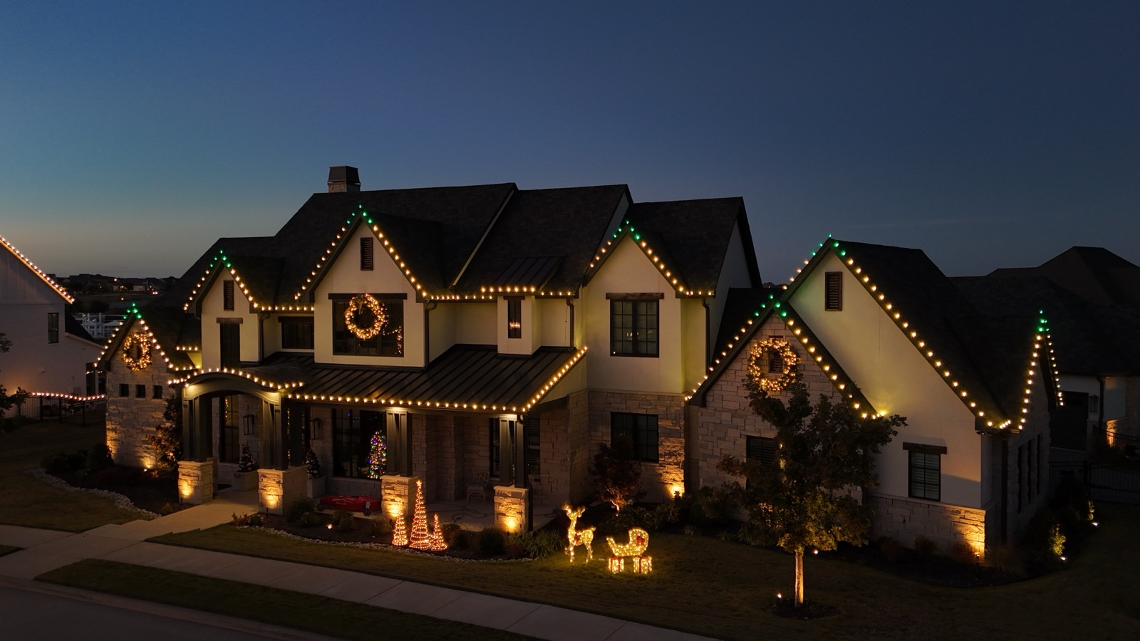 Two-story stone and stucco home with warm white roofline lighting, lit wreaths, and yard displays at dusk