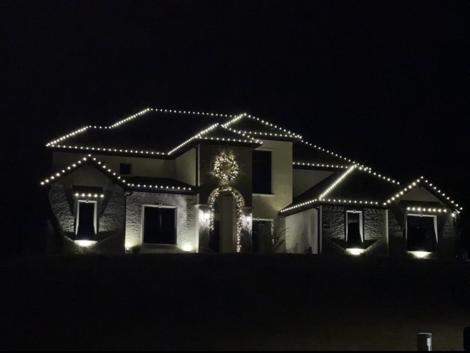 Stucco two-story home with warm white roofline lighting and a lit wreath and garland entrance arch